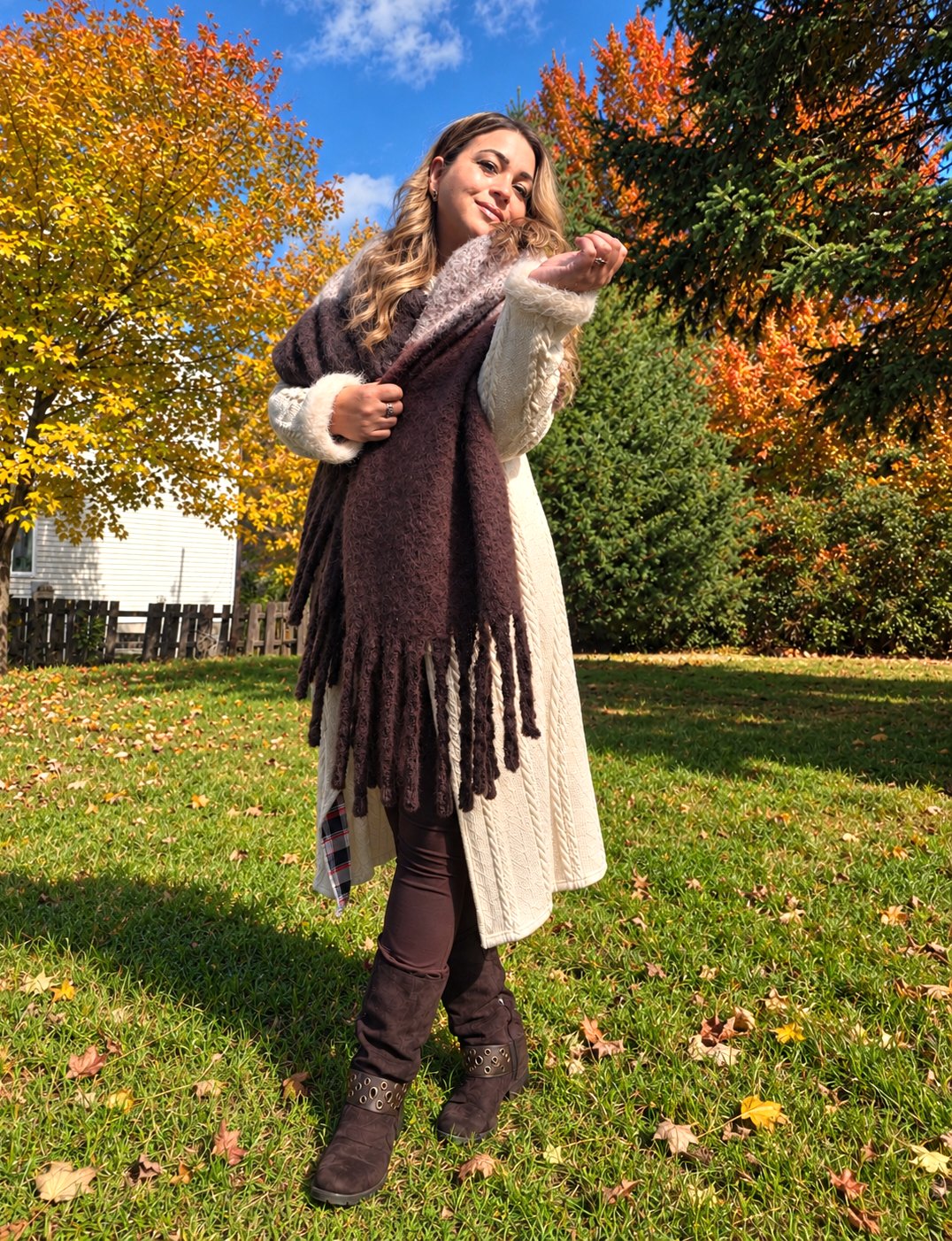 Cindy Roy en plein corps dans son jardin en automne — long cardigan crème, grand foulard brun à franges et bottes brunes — envoyant un baiser vers la caméra, entourée d’arbres aux feuilles dorées et orangées