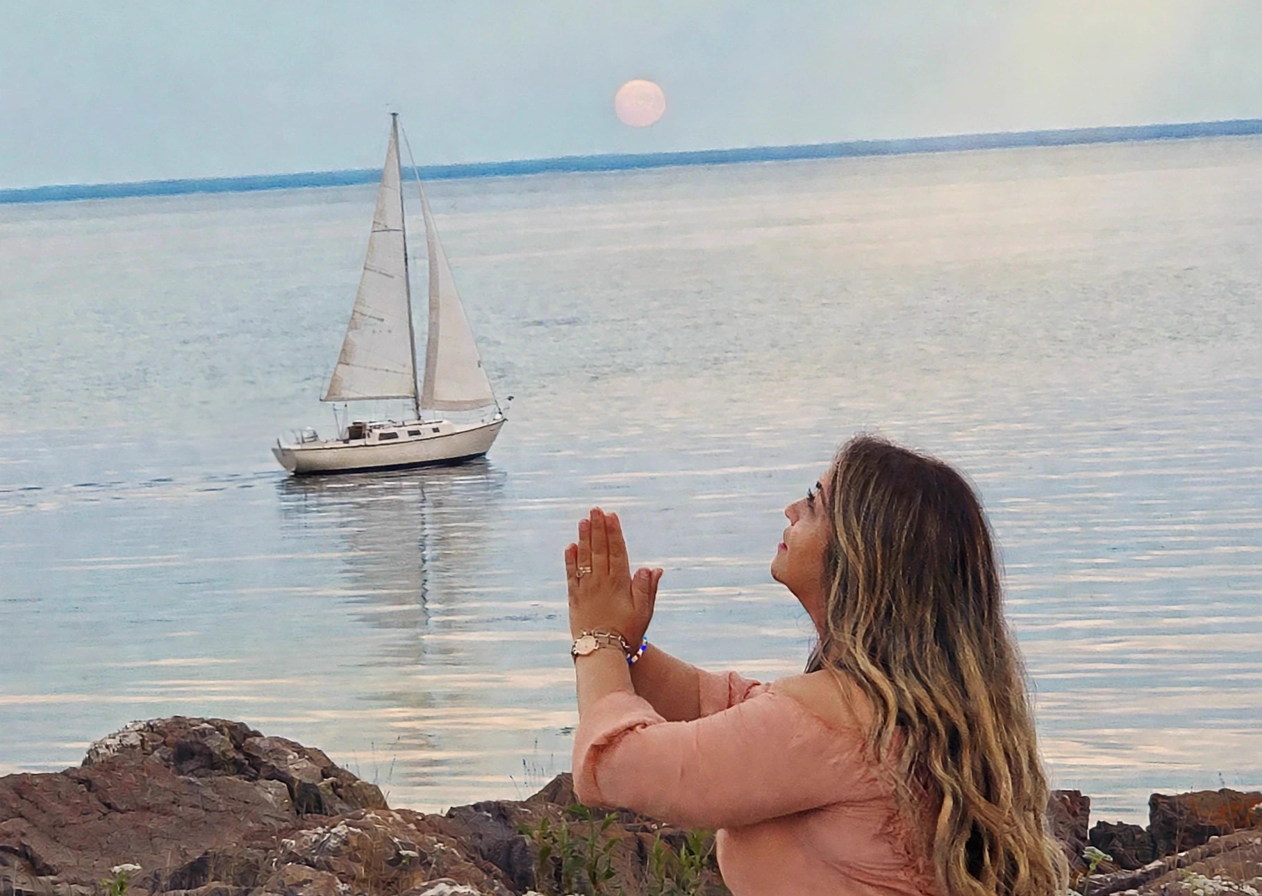 Cindy Roy, en blouse pêche, les mains jointes devant la mer au crépuscule — un voilier navigue sous la pleine lune rose