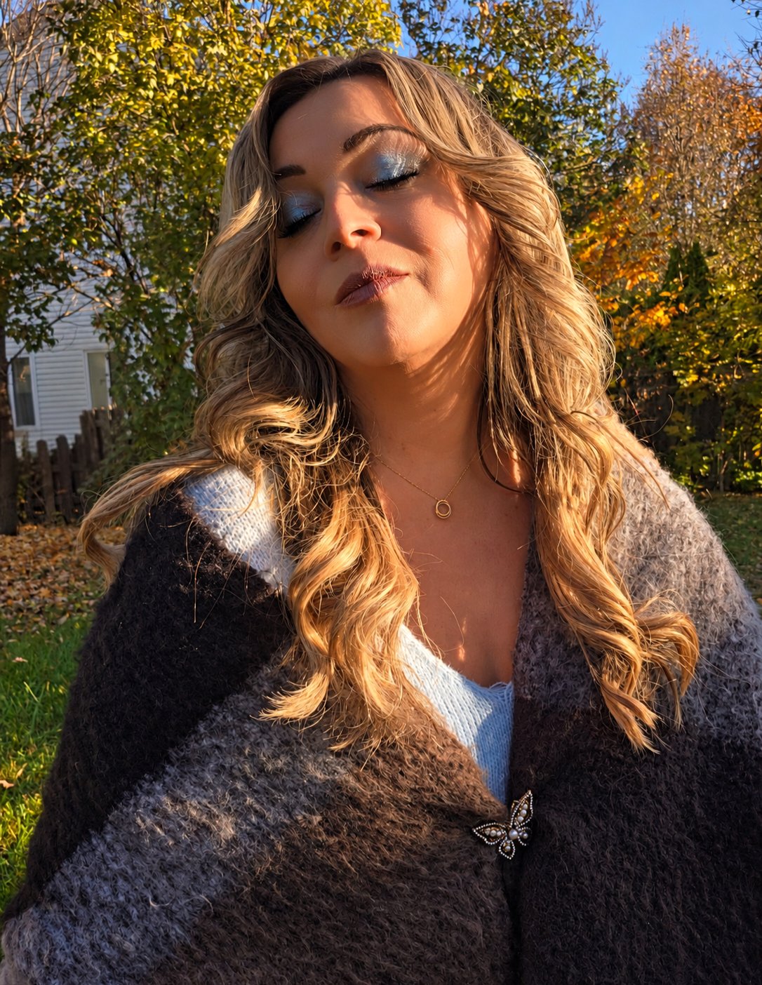 Cindy Roy les yeux fermés, maquillage bleu, portant son châle tricoté brun et gris et sa broche papillon — pose artistique dans la lumière dorée