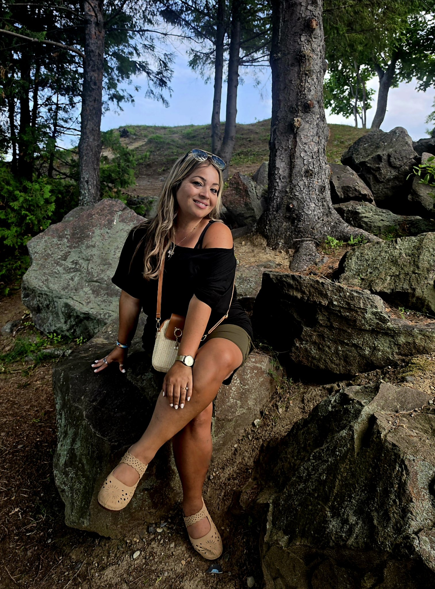 Cindy Roy assise sur un rocher sous les pins, souriante, les lunettes de soleil sur la tête