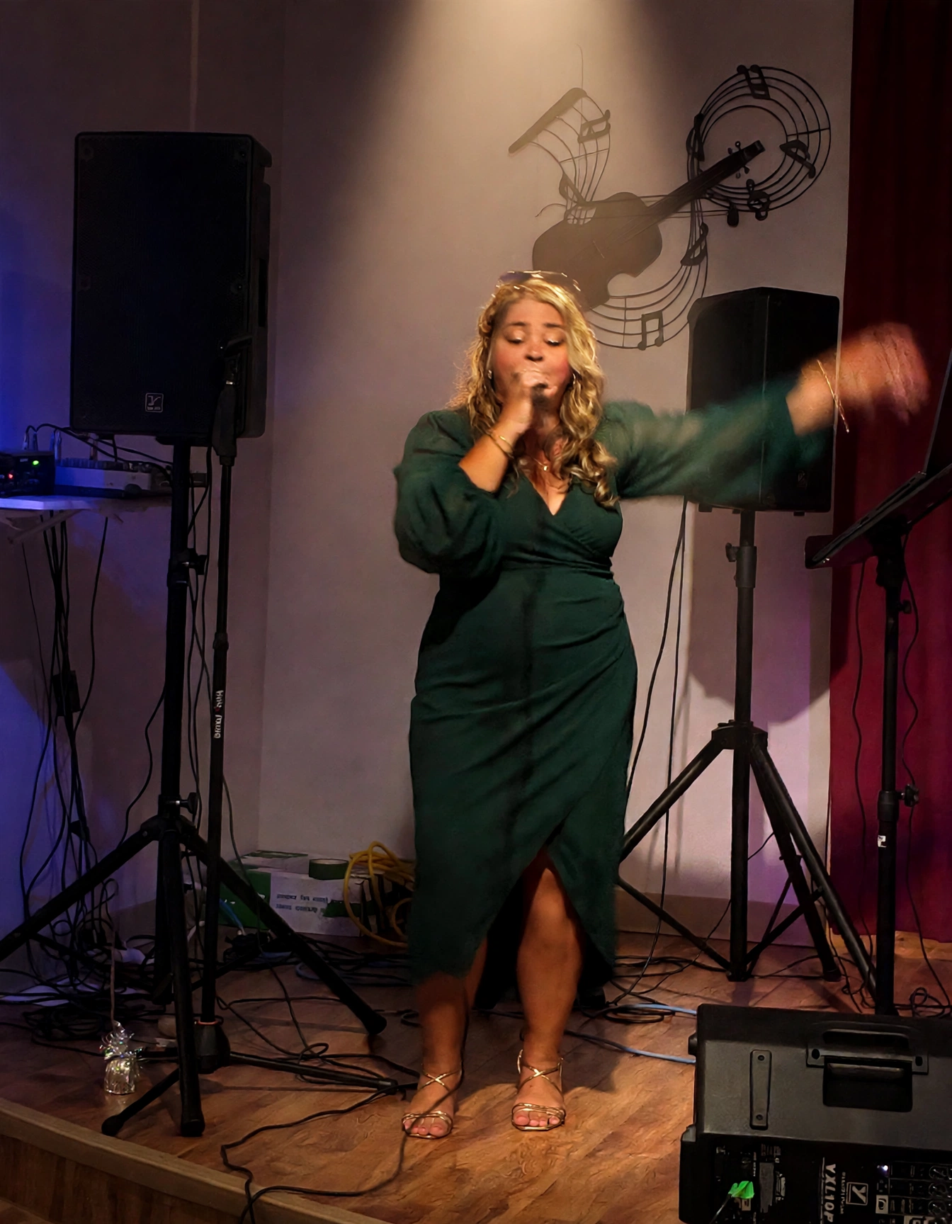 Cindy Roy en spectacle le soir, en robe verte et talons dorés, le micro à la bouche sous un spot chaud, un bras tendu vers le public
