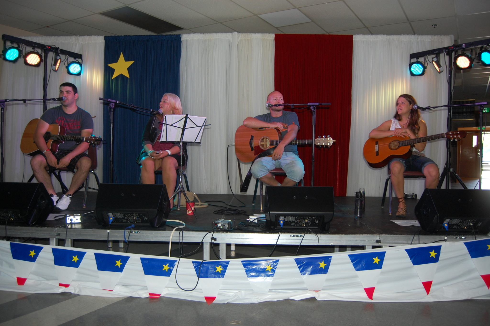 Le cercle d’auteurs-compositeurs-interprètes sur scène aux Festivités acadiennes de Beresford, en août 2015 — de gauche à droite : Louis Noël, Cindy Roy, Stéphane Hébert et Madiane Michaud, devant un grand drapeau acadien