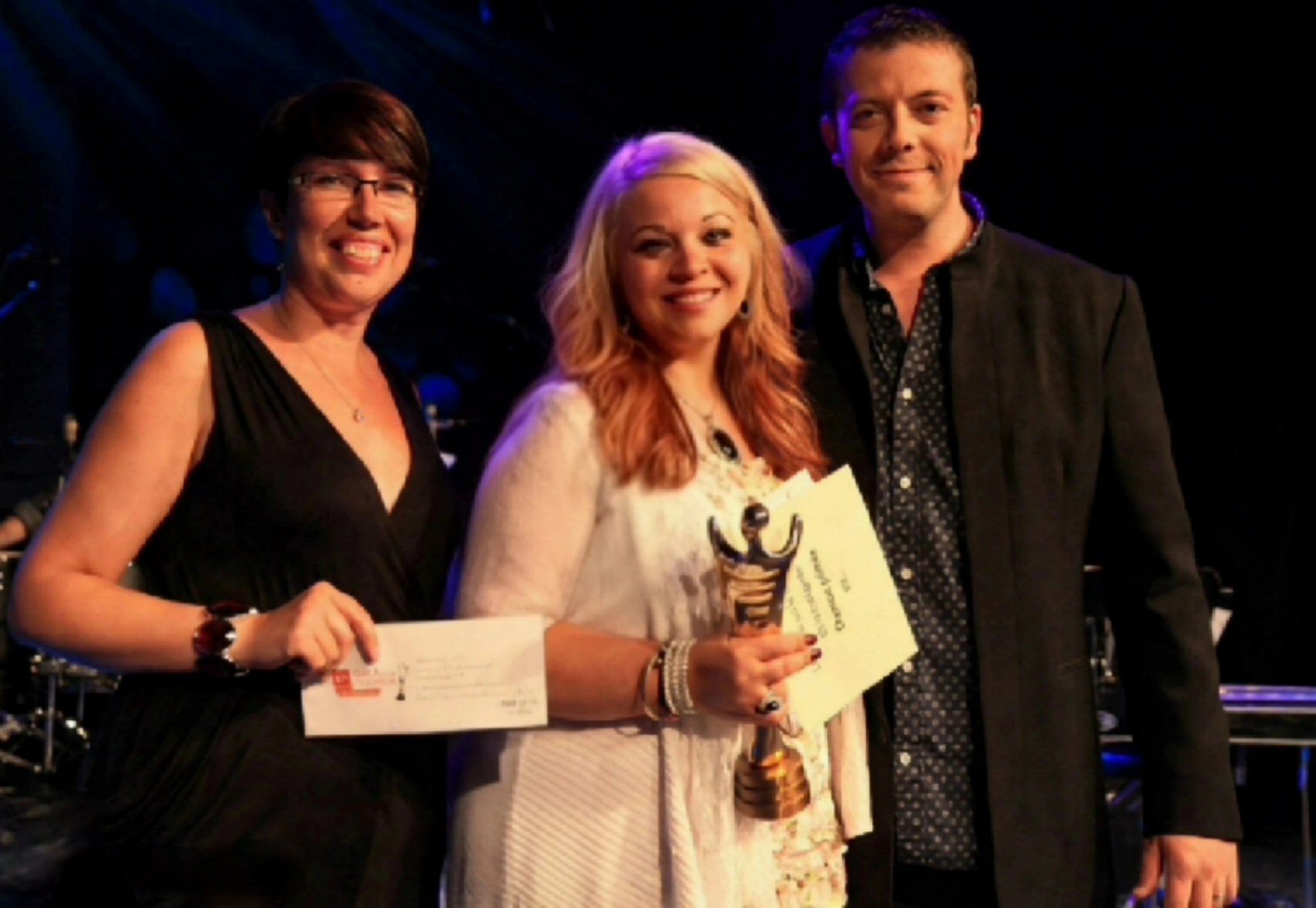Cindy Roy sur scène, radieuse, tenant son trophée et son certificat, entourée de deux présentateurs lors de sa victoire au 47e Gala de la chanson de Caraquet en 2015