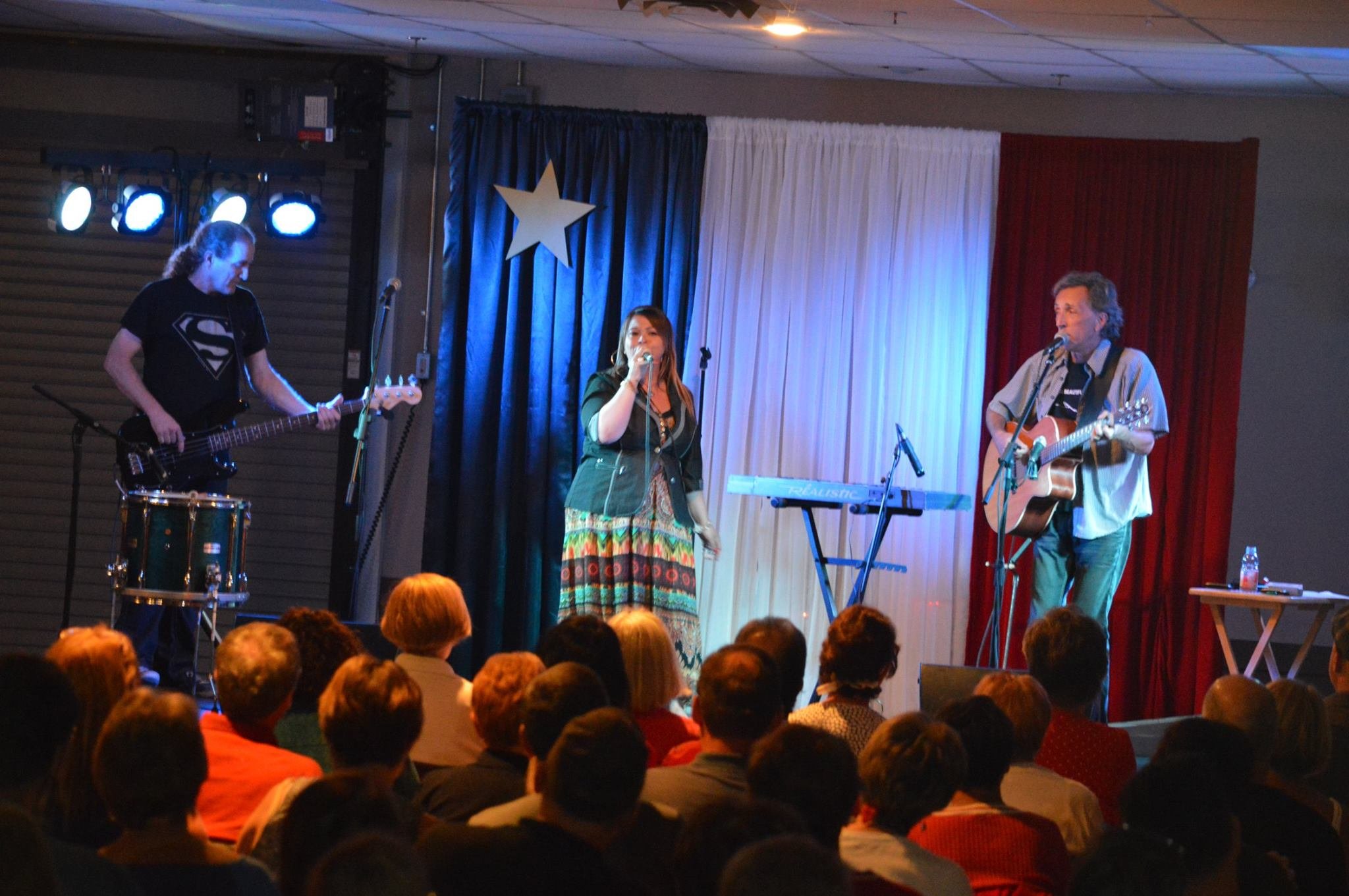 Cindy Roy au micro sur scène avec Michel Thériault à la guitare et son bassiste, devant un rideau aux couleurs acadiennes — Beresford, 15 août 2016